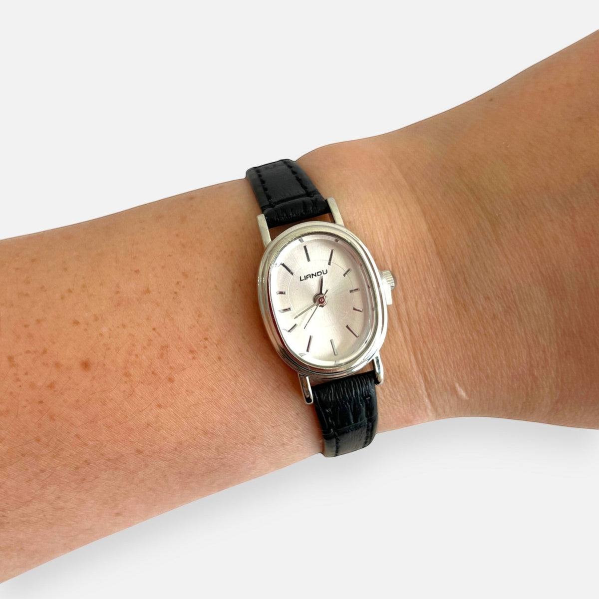 Reloj Hv Hermanas Villegas con caja ovalada plateada, dial satinado y correa de cuero vegano negro en la muñeca