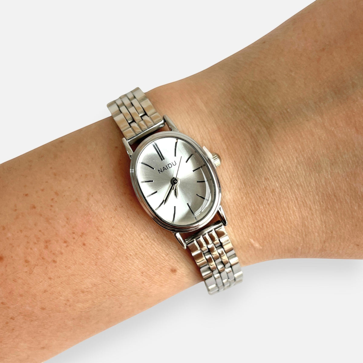 Reloj analógico Hv Hermanas Villegas con caja ovalada plateada, dial satinado y correa de acero inoxidable en muñeca