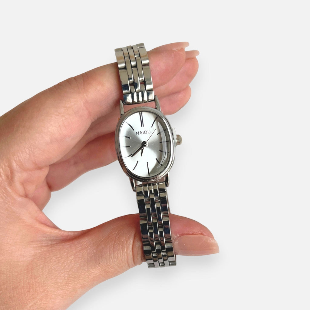 Reloj analógico Hv Hermanas Villegas con caja ovalada plateada, dial satinado y correa de acero inoxidable.