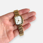 Reloj Hv Hermanas Villegas rectangular dorado con correa de eslabones y dial claro, sostenido en una mano.