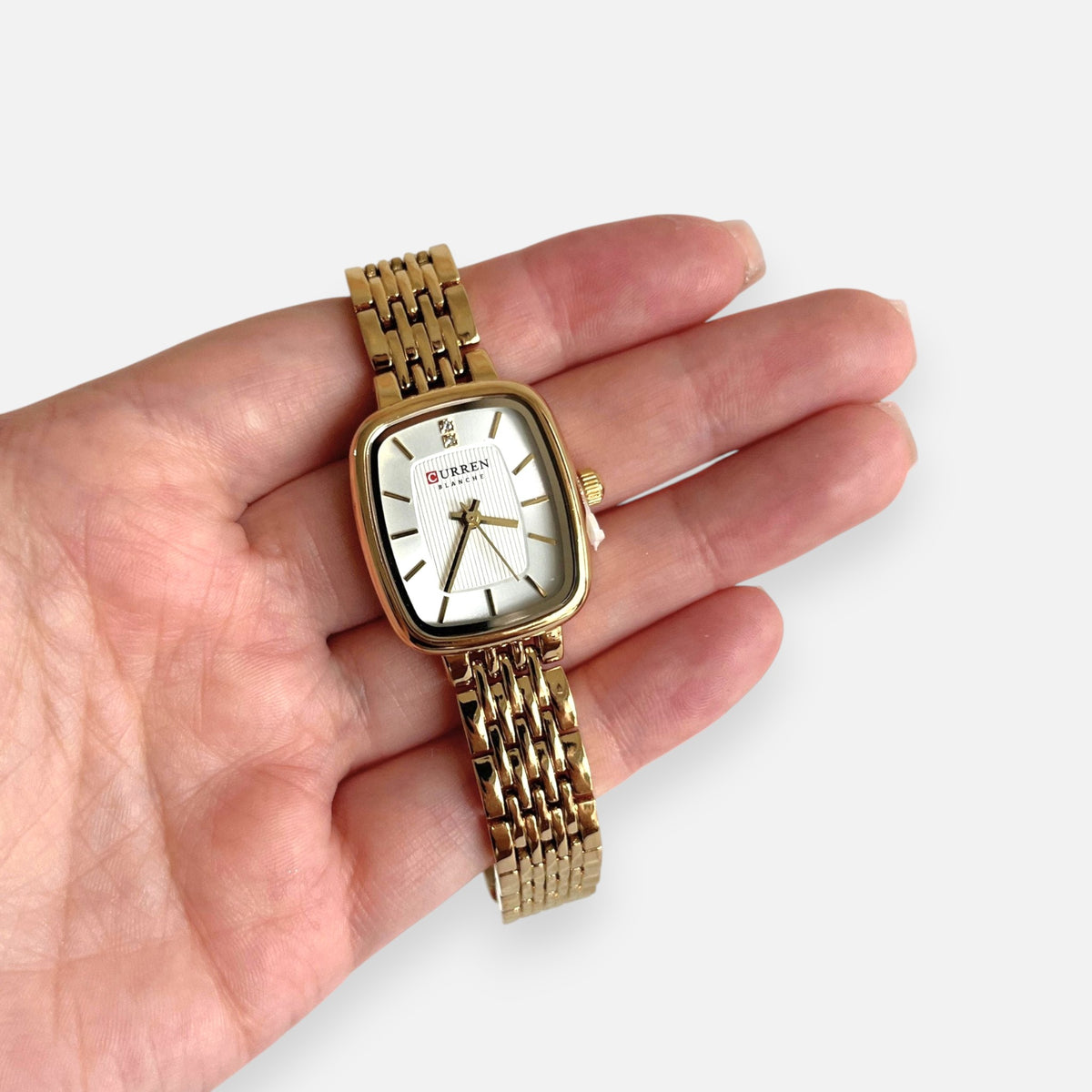 Reloj Hv Hermanas Villegas rectangular dorado con correa de eslabones y dial claro, sostenido en una mano.